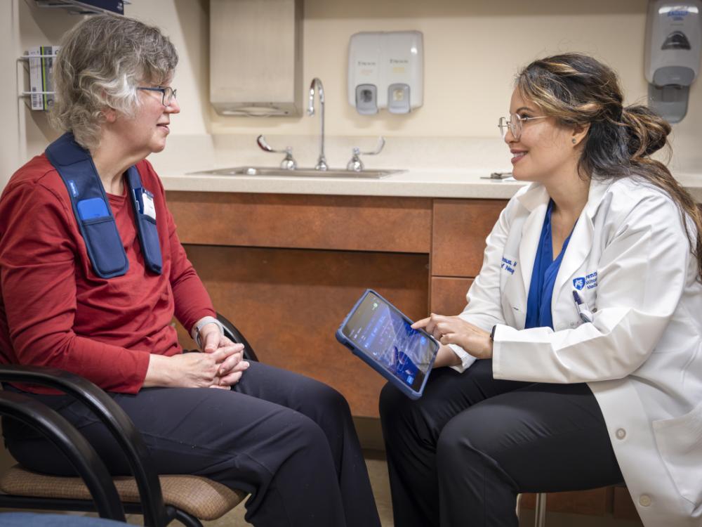 A physician sits next to a patient in a clinic room. The physician holds a tablet computer as she smiles and looks at the patient.