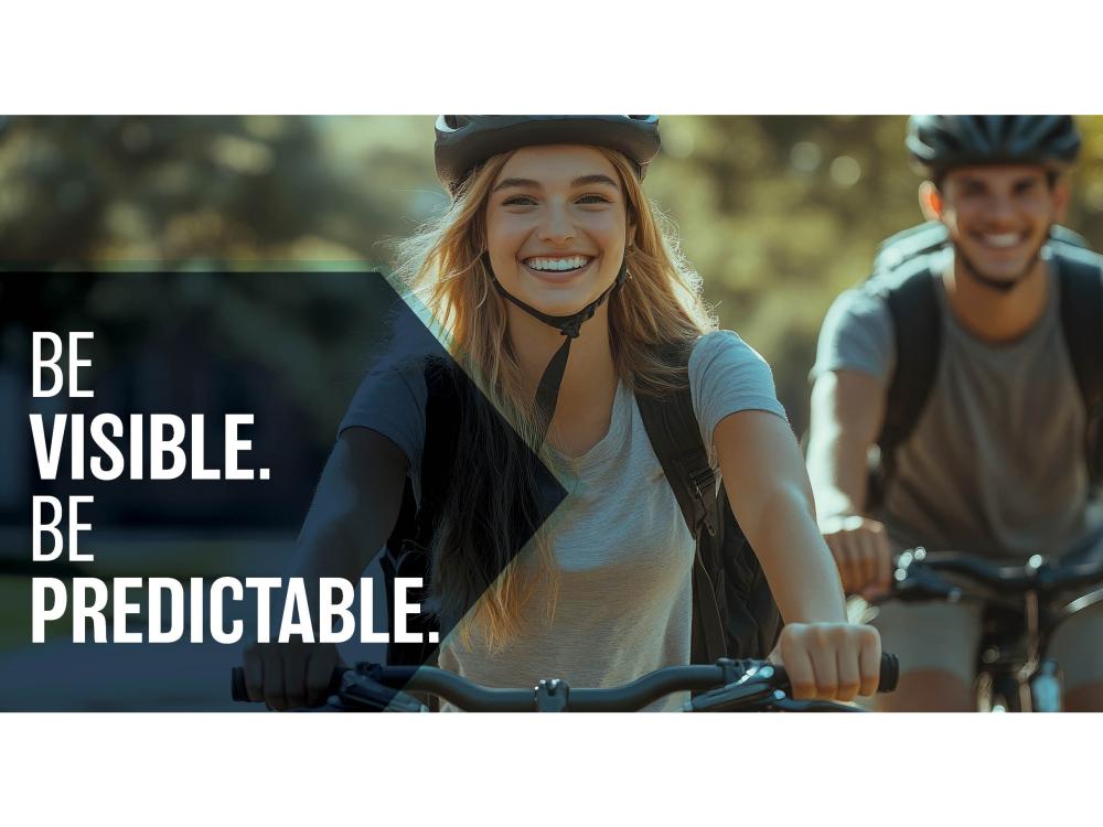 Two college students riding bikes, "Be Visible. Be Predictable"