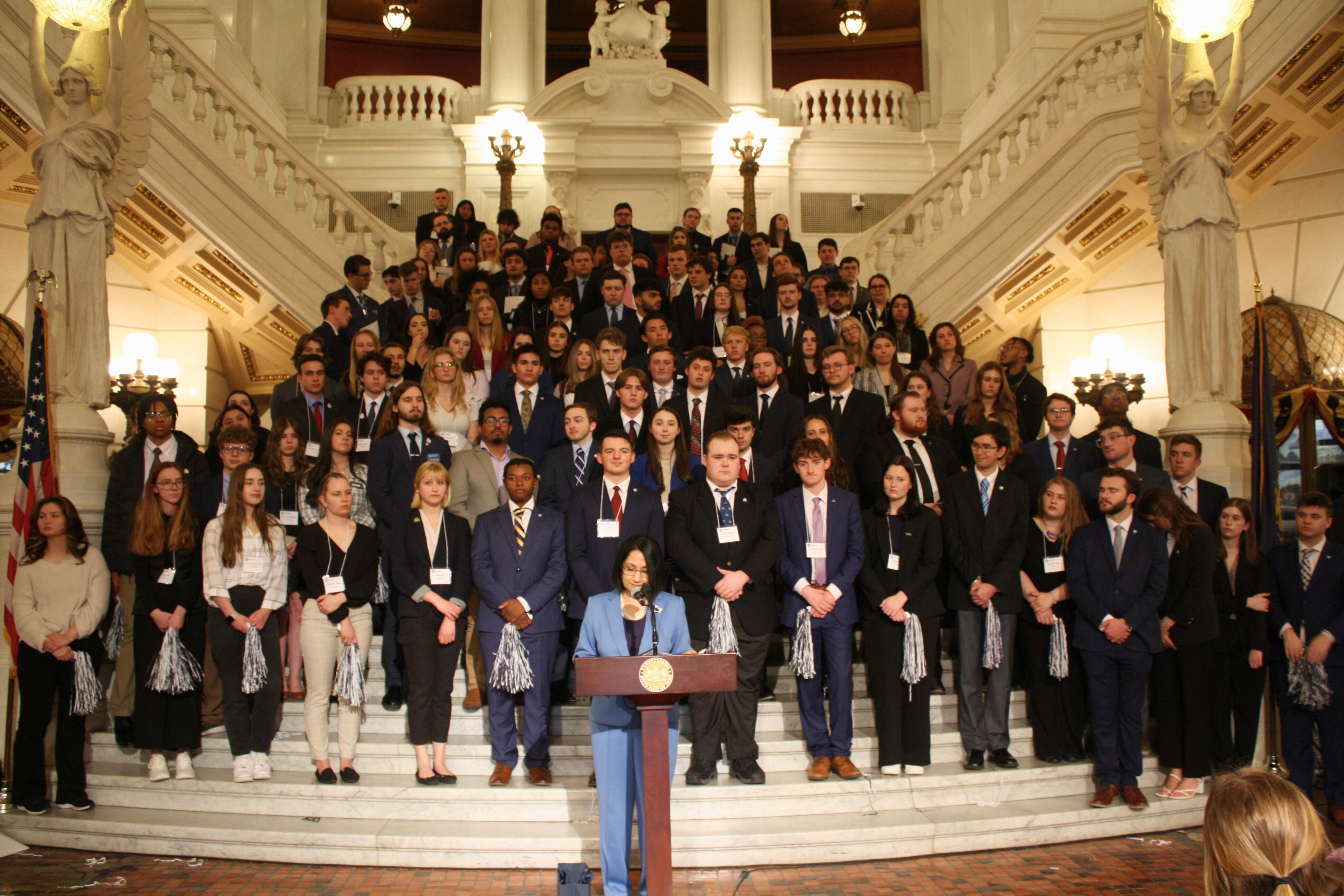 A large group of students in business attire stands on a grand staircase, listening to a speaker at a podium in a historic government building