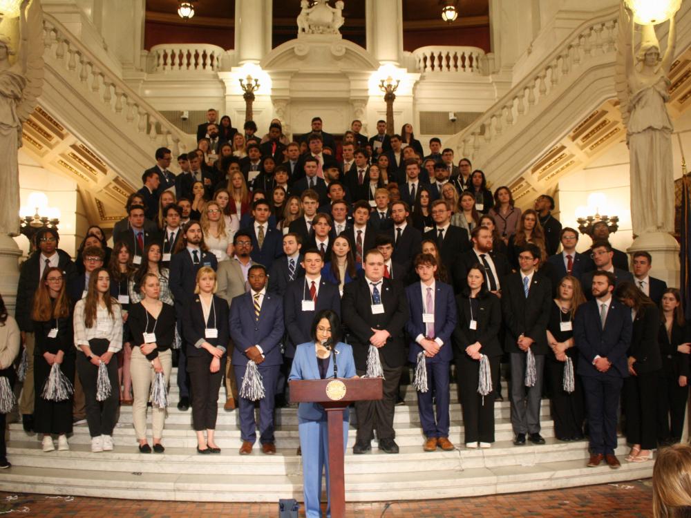 A large group of students in business attire stands on a grand staircase, listening to a speaker at a podium in a historic government building