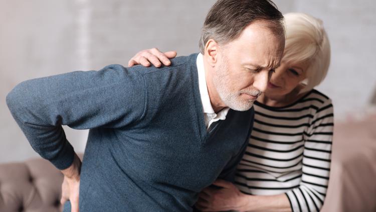 An older man looking in pain and holding his lower back being support by an older woman