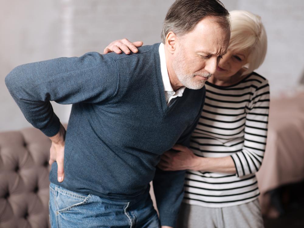 An older man looking in pain and holding his lower back being support by an older woman