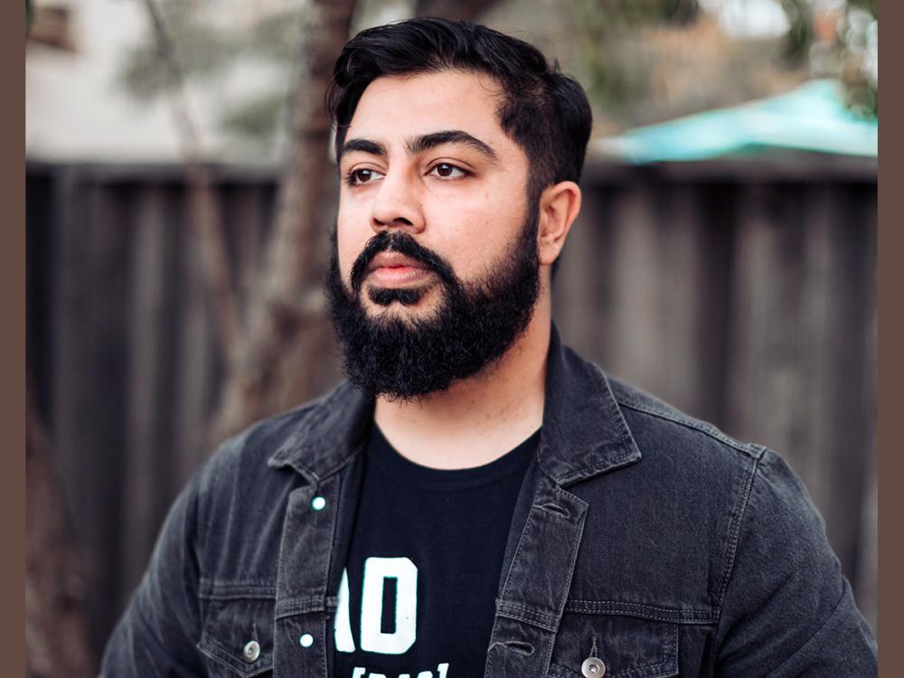 Photo of fiction writer Jamil Jan Kochai standing outside in front of brown picket fence