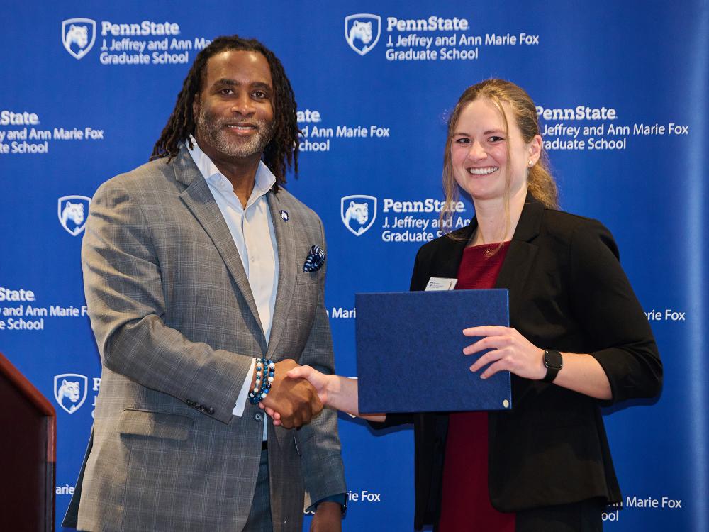 Tricia Hart stands on stage holding a certificate while shaking hands with Dean Levon Esters