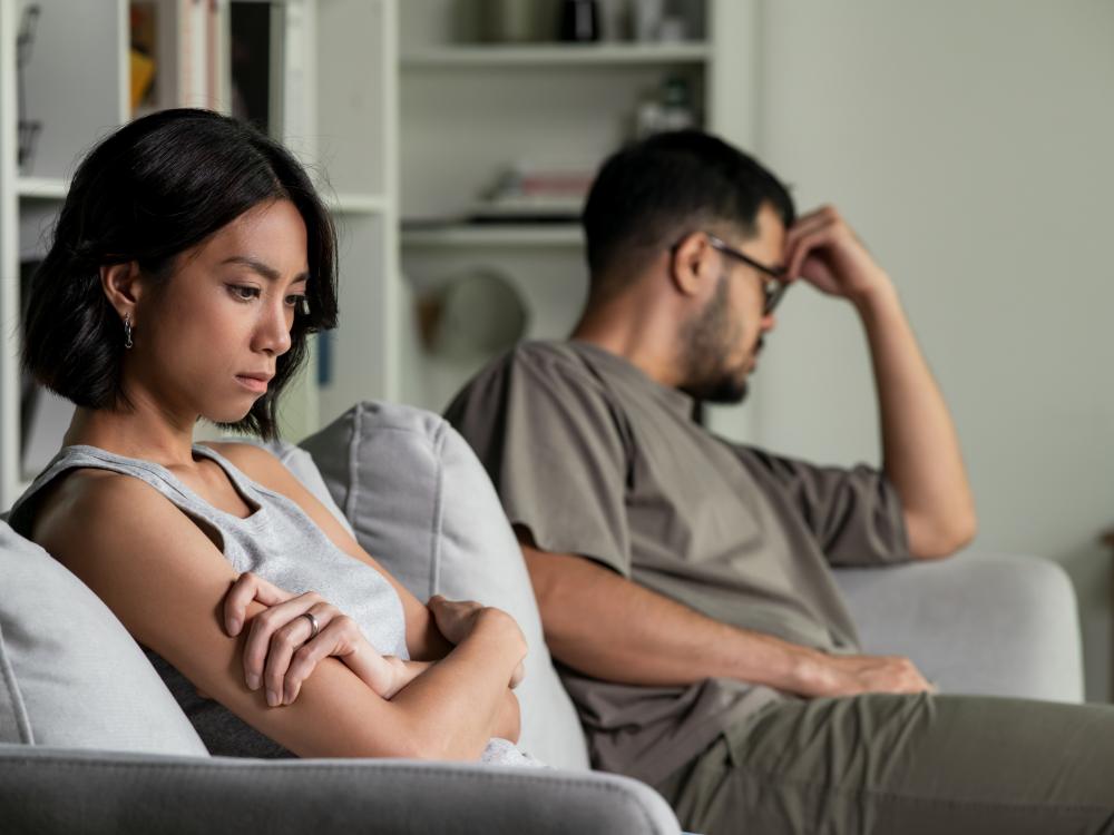 Two people sit on opposite ends of a couch with unhappy expressions, looking away from each other