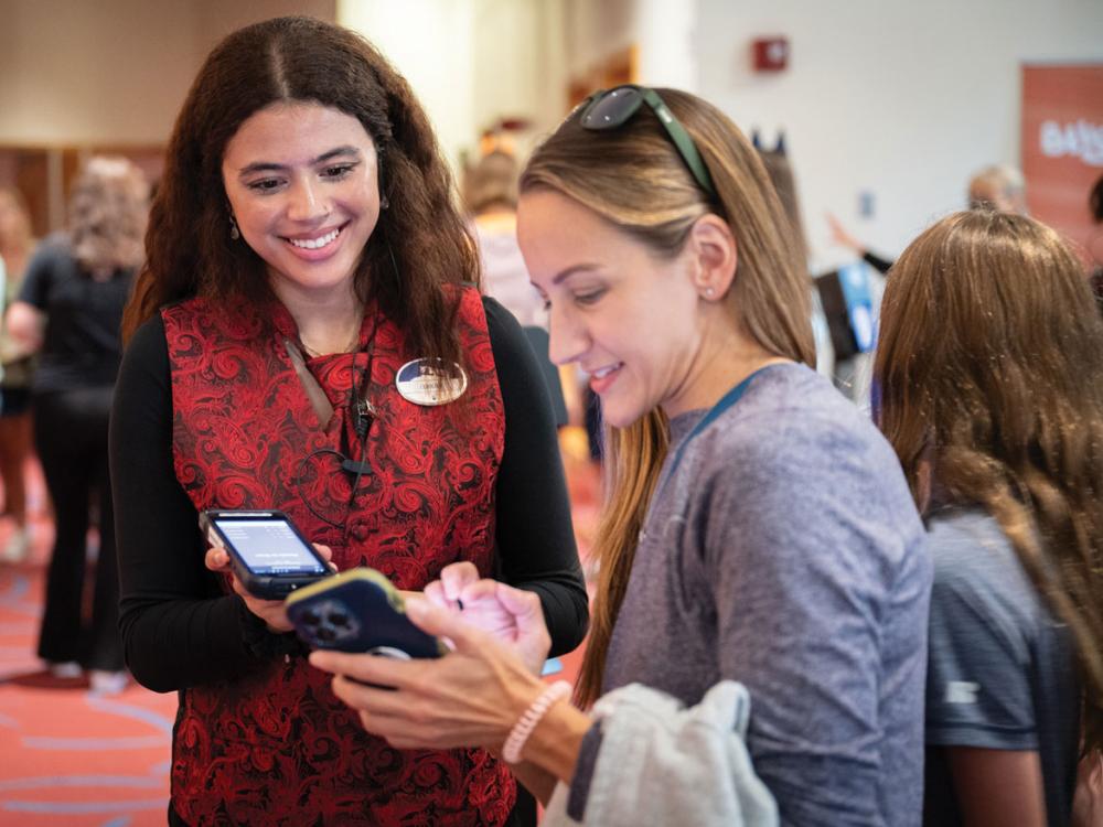 A young woman assists another woman with accessing tickets on her mobile phone.