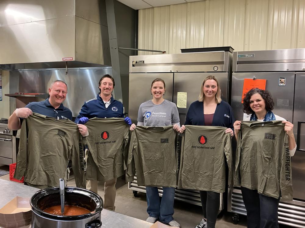 Five people hold long sleeve, green T-shirts in a kitchen.