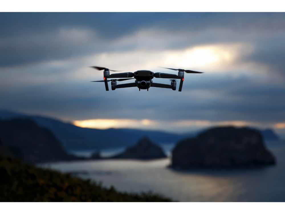 A drone flies in the sky over a blurred-out body of water.